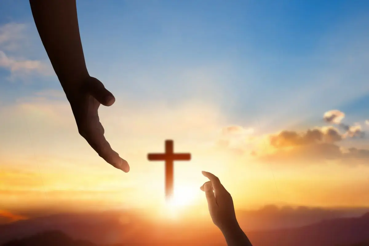 Two hands, symbolizing a diverse church congregation, reach toward each other with a cross in the distance against a sunset sky.