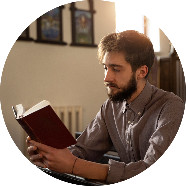 A man with a beard and brown hair sits indoors, reading a book about intercultural church services. Clad in a gray shirt, he is surrounded by framed artwork and soft lighting, creating an inviting atmosphere for reflection.