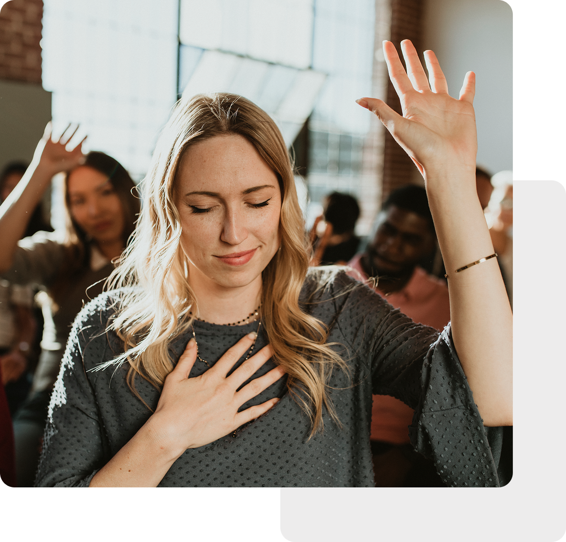 In a sunlit room, a woman with eyes closed raises her hand amidst a diverse church congregation gathered for an uplifting event.