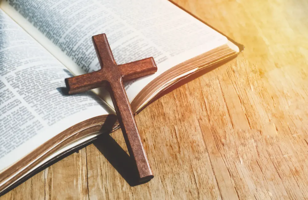 An open Bible lies on a wooden surface with a wooden cross resting on its pages in sunlight.