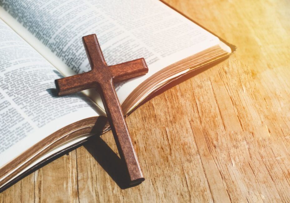 An open Bible lies on a wooden surface with a wooden cross resting on its pages in sunlight.